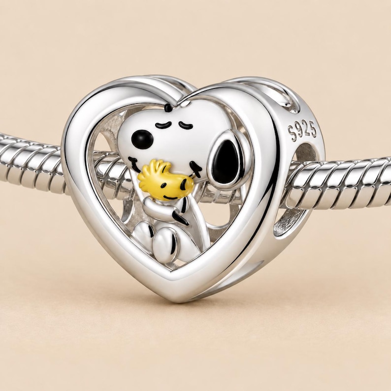 May include: A silver heart-shaped charm bead featuring a white Snoopy character holding a yellow Woodstock figure. The charm is threaded onto a silver snake chain bracelet. The side of the heart is engraved with the text S925, indicating sterling silver.