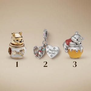 Puede incluir: Tres dijes de metal con personajes de Winnie the Pooh. El dije 1 es un oso dorado en una maceta blanca con una abeja. El dije 2 es un colgante de plata en forma de corazón con el texto "Happy partner". El dije 3 es un oso plateado sobre un tarro de miel.