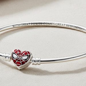 Op de afbeelding: Zilverkleurige armband met een hartvormige bedel met een rood en wit Spider-Man-ontwerp. De armband is voorzien van de tekst "WITH GREAT POWER COMES GREAT RESPONSIBILITY". Een stijlvol accessoire voor superheldenfans.