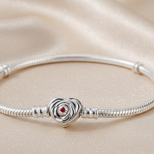 May include: A silver bracelet with a heart-shaped charm. The charm features a rose design with a small red gemstone in the center. The bracelet has a snake chain design and is displayed on a beige satin surface.