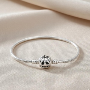 May include: A silver snake chain bracelet featuring a central charm shaped like a pumpkin with carved facial features. The pumpkin charm has dark recessed areas for the eyes and mouth, contrasting with the polished silver finish of the bracelet.