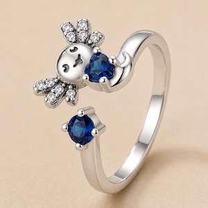 Puede incluir: Un anillo abierto de tono plateado con un diseño de ajolote con piedras transparentes en las branquias. El ajolote sostiene una piedra azul oscuro en forma de corazón, mientras que el otro extremo tiene una piedra redonda azul oscuro engastada.