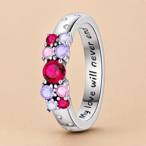 Puede incluir: Un anillo de tono plateado con un grupo de piedras de corte redondo en colores rojo, rosa y morado claro. La banda tiene grabado el texto My love will never end e incluye pequeñas piedras transparentes incrustadas en la superficie exterior.