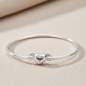 May include: A silver bangle bracelet with a heart-shaped charm. The charm has the words "Always by your side" engraved on it, with small wings on either side. The bracelet is made of a shiny, smooth metal.