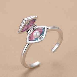 Puede incluir: Un anillo abierto de tono plateado con un diseño de concha marina articulada. El interior de la concha está esmaltado con tonos degradados en rosa y azul, conteniendo una perla esférica blanca y una pequeña piedra rectangular transparente.