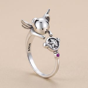 May include: A silver open ring featuring a small winged creature with a rounded head and a small mouth. The ring band is stamped with S925 and terminates in a circular charm containing a five-pointed star and a small, round, magenta-coloured stone.