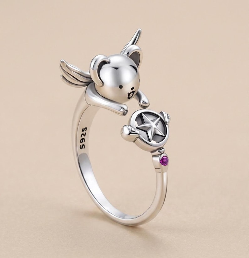 May include: A silver open ring featuring a small winged creature with a rounded head and a small mouth. The ring band is stamped with S925 and terminates in a circular charm containing a five-pointed star and a small, round, magenta-coloured stone.
