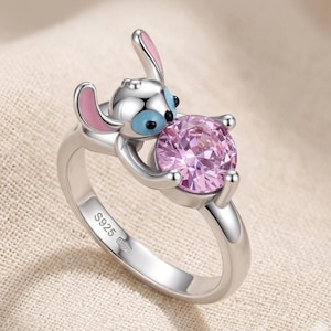 May include: A silver ring featuring a cartoon character with blue eyes and pink ears, holding a large, round, pink gemstone. The ring is stamped with "S925".
