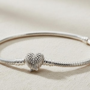 Puede incluir: Un brazalete plateado con un cierre en forma de corazón. El brazalete tiene un diseño de cadena de serpiente y un cierre en forma de corazón con un patrón similar a una pluma. El brazalete está sobre una superficie neutra.