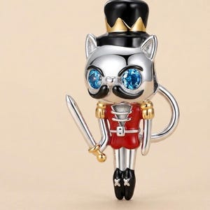 May include: A silver-toned metal charm shaped like a cat dressed as a nutcracker. The cat wears a black top hat with a gold crown, has bright blue gemstone eyes, a black mustache, a red uniform jacket, and holds a silver sword in one hand.