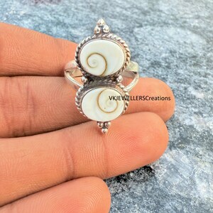 May include: A silver ring with two oval, cream-coloured stones, each with a spiral pattern. The stones are set in a decorative silver frame with a beaded edge. The ring is on a hand.