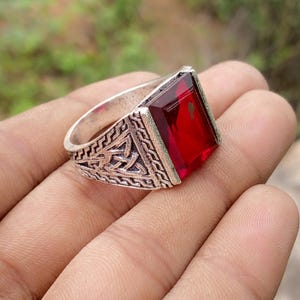 Puede incluir: Un anillo de metal plateado con una gran piedra preciosa de color rojo intenso, rectangular y facetada, engastada en el centro. La banda muestra un patrón intrincado, elevado y texturizado en los lados, dando un acabado detallado al metal.