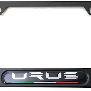 May include: Black license plate frame with the word "URUS" in white lettering. The frame has a rectangular shape with rounded corners and mounting holes. A small Italian flag is at the bottom.