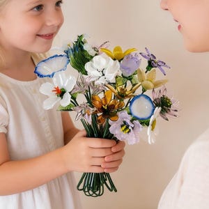May include: A young girl in a white dress presents a colorful bouquet of glass flowers to a woman. The bouquet includes white, blue, yellow, and purple flowers, held together with green wire. The woman is wearing a light-colored top.