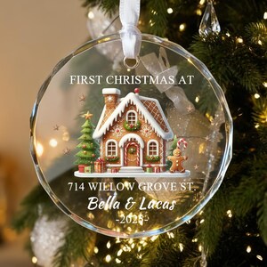 May include: A clear, round Christmas ornament with a gingerbread house design. The ornament has the text "FIRST CHRISTMAS AT" and the address "714 WILLOW GROVE ST." with names and a year. It hangs from a white ribbon.