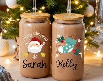 May include: Two glass tumblers with bamboo lids and straws, featuring Christmas-themed designs. One has a Santa Claus graphic and the text "Merry Christmas Sarah", the other has a stocking graphic and the name "Kelly". A small Christmas tree is in the background.