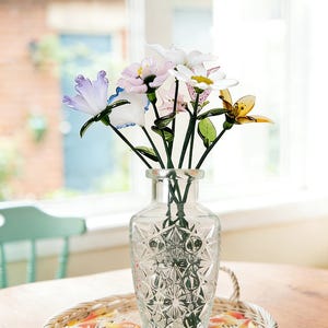 May include: A clear glass vase with a geometric pattern holds a bouquet of colorful glass flowers. The flowers include white, pink, yellow, and blue petals with green stems. The vase sits on a woven tray with a decorative border.
