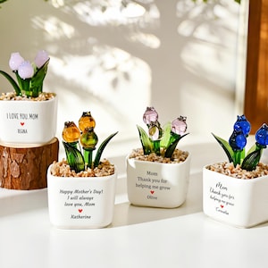 Puede incluir: Cuatro macetas de cerámica blanca con flores de tulipanes de cristal de varios colores. Cada maceta tiene un mensaje y un nombre. Los tulipanes son en tonos rosa, amarillo y azul, con hojas verdes.