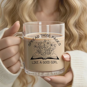 Pode incluir: Uma caneca de vidro transparente cheia de uma bebida cremosa. A caneca apresenta um gráfico preto de um livro aberto com flores e o texto "SPREAD THOSE PAGES LIKE A GOOD GIRL". A caneca tem uma alça e é segurada por uma pessoa.