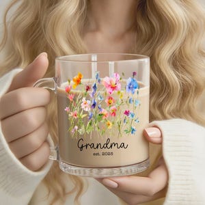 Op de afbeelding: Een heldere glazen mok gevuld met een lichtbruine vloeistof, met een bloemenontwerp van roze, blauwe, oranje en gele bloemen. De mok is bedrukt met "Grandma" en het jaar "est. 2025" in zwarte letters.
