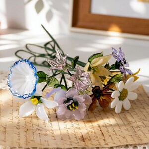 May include: A colorful glass flower bouquet featuring a variety of blooms in white, blue, yellow, and purple. The flowers are arranged with green stems and leaves, resting on a vintage paper with handwritten text. A red strawberry is visible in the background.