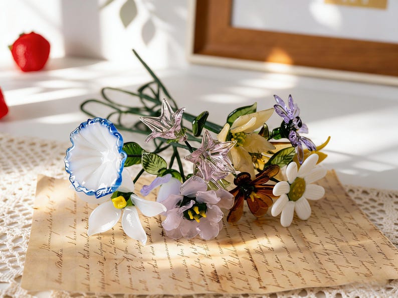 May include: A colorful glass flower bouquet featuring a variety of blooms in white, blue, yellow, and purple. The flowers are arranged with green stems and leaves, resting on a vintage paper with handwritten text. A red strawberry is visible in the background.