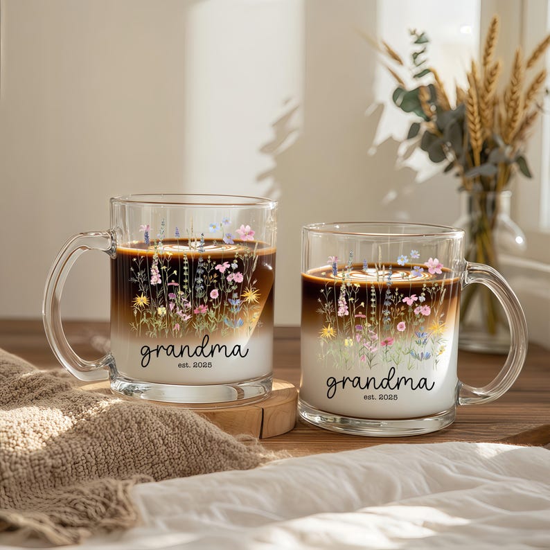 Puede incluir: Dos tazas de vidrio transparente llenas de una bebida de caf&eacute; en capas. Cada taza presenta un dise&ntilde;o floral con la palabra "grandma" y el a&ntilde;o "est. 2025" en escritura negra. Las tazas tienen un asa y est&aacute;n sobre una superficie de madera.