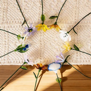 May include: A collection of colorful glass flowers with green stems and leaves. The flowers are in various colors, including yellow, white, blue, orange, and purple. The flowers are arranged on a cream-colored crocheted surface.