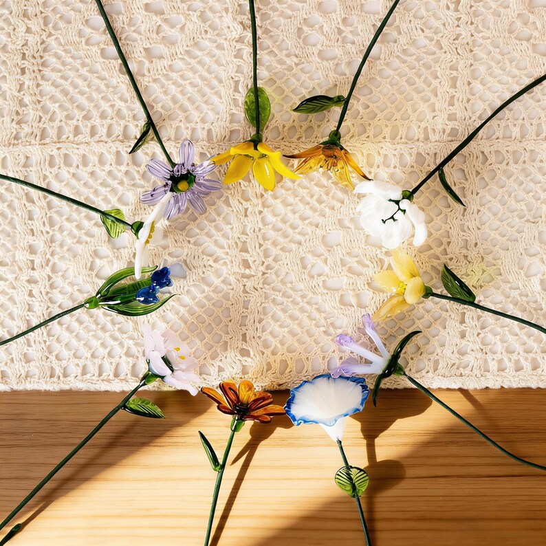 May include: A collection of colorful glass flowers with green stems and leaves. The flowers are in various colors, including yellow, white, blue, orange, and purple. The flowers are arranged on a cream-colored crocheted surface.