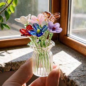 Puede incluir: Un pequeño jarrón de cristal lleno de coloridas flores de cristal. Las flores son de varios colores, incluyendo rojo, azul, rosa, blanco y ámbar. El jarrón se sostiene en una mano, con una ventana y vegetación en el fondo.