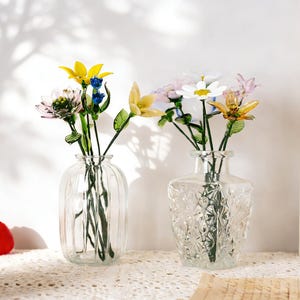May include: Two clear glass vases with colorful glass flowers. One vase is ribbed, the other is square with a textured pattern. The flowers include yellow, white, pink, and blue blooms with green stems and leaves. The vases sit on a white lace tablecloth.