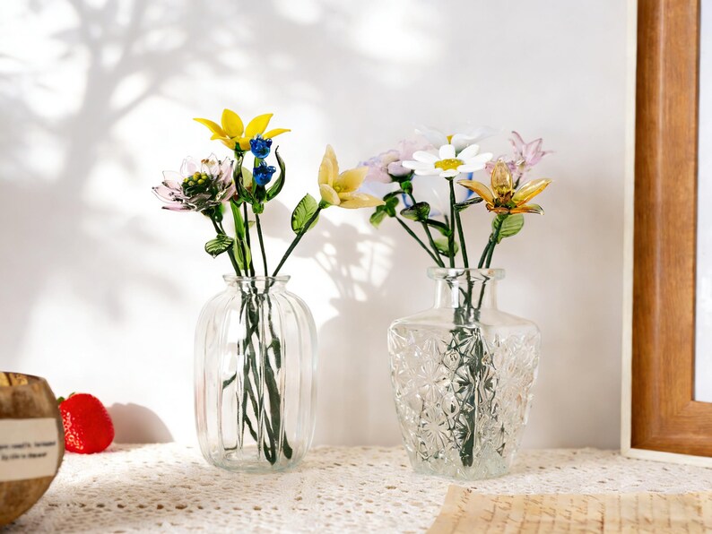 May include: Two clear glass vases with colorful glass flowers. One vase is ribbed, the other is square with a textured pattern. The flowers include yellow, white, pink, and blue blooms with green stems and leaves. The vases sit on a white lace tablecloth.