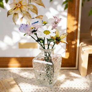 May include: A clear, faceted glass vase holds a small bouquet of colorful artificial flowers. The flowers include white daisies, purple blooms, and yellow-green accents. The vase sits on a lace tablecloth, with a framed floral print in the background.