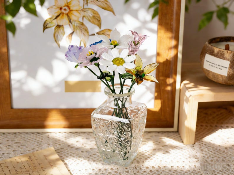 May include: A clear, faceted glass vase holds a small bouquet of colorful artificial flowers. The flowers include white daisies, purple blooms, and yellow-green accents. The vase sits on a lace tablecloth, with a framed floral print in the background.