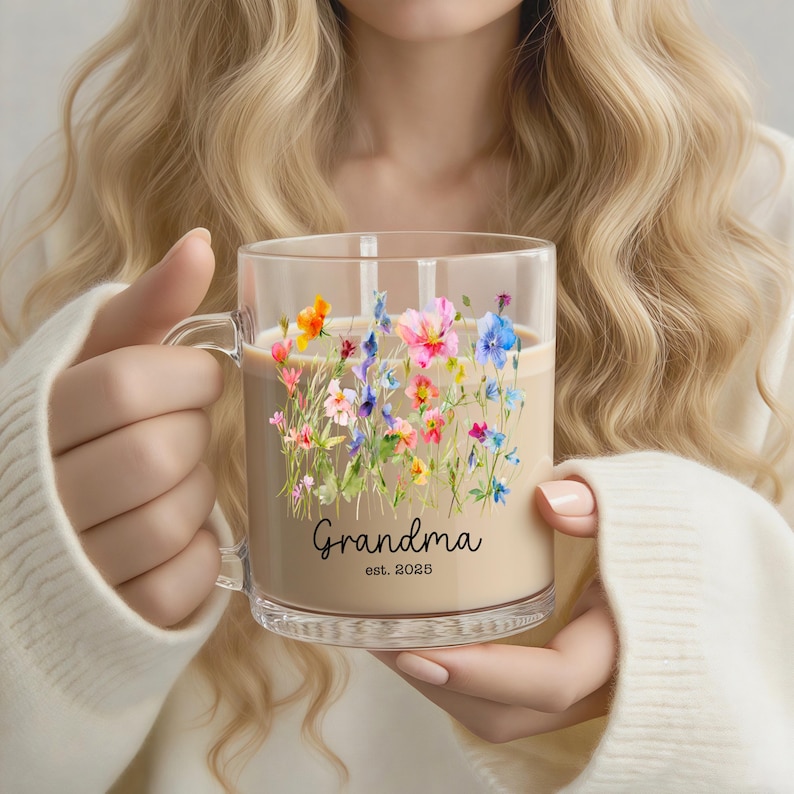 Puede incluir: Taza de vidrio transparente llena de un l&iacute;quido marr&oacute;n claro, con un dise&ntilde;o floral de flores rosas, azules, naranjas y amarillas. La palabra "Grandma" y el a&ntilde;o "est. 2025" est&aacute;n impresos en negro en la taza.