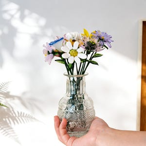 May include: A clear glass vase with a textured pattern holds a bouquet of colorful flowers. The flowers include white daisies, purple, yellow, and pink blooms with green stems. The vase is held by a hand, with a white wall and a wooden framed picture in the background.