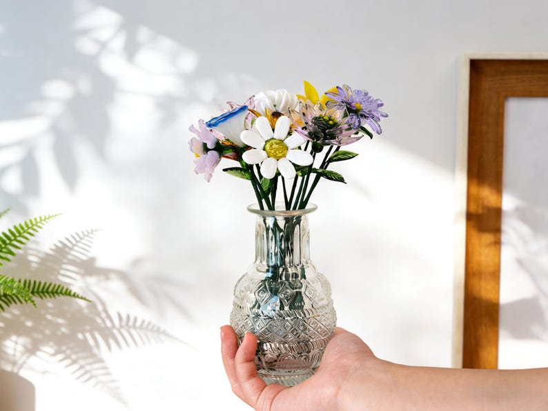 May include: A clear glass vase with a textured pattern holds a bouquet of colorful flowers. The flowers include white daisies, purple, yellow, and pink blooms with green stems. The vase is held by a hand, with a white wall and a wooden framed picture in the background.