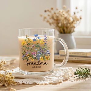 Puede incluir: Una taza de vidrio transparente llena de una bebida de color claro. La taza est&aacute; decorada con un dise&ntilde;o floral en tonos amarillos, azules, morados y rosas. La palabra "Grandma" y el a&ntilde;o "est. 2025" est&aacute;n impresos en la taza.