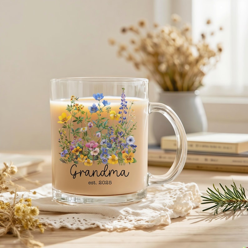 Puede incluir: Una taza de vidrio transparente llena de una bebida de color claro. La taza est&aacute; decorada con un dise&ntilde;o floral en tonos amarillos, azules, morados y rosas. La palabra "Grandma" y el a&ntilde;o "est. 2025" est&aacute;n impresos en la taza.