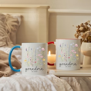 Puede incluir: Dos tazas de cer&aacute;mica blancas con dise&ntilde;os florales y la palabra "grandma" en escritura negra. Una taza tiene un asa azul, la otra un asa roja. El texto "est. 2025" est&aacute; debajo de la palabra "grandma".