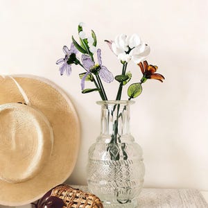 May include: A clear glass vase with a textured design holds colorful glass flowers. The flowers include white, purple, and orange blooms with green stems and leaves. A straw hat and woven basket are in the background.