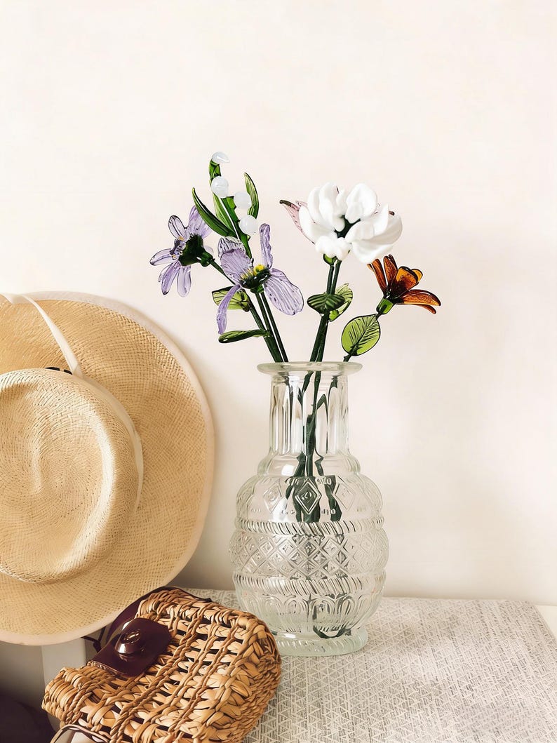 May include: A clear glass vase with a textured design holds colorful glass flowers. The flowers include white, purple, and orange blooms with green stems and leaves. A straw hat and woven basket are in the background.