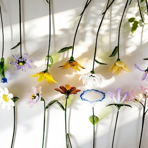 May include: A collection of colorful glass flowers with long green stems. The flowers are in various colors, including yellow, white, purple, and orange. The stems are arranged vertically against a white background.