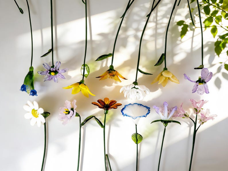 May include: A collection of colorful glass flowers with long green stems. The flowers are in various colors, including yellow, white, purple, and orange. The stems are arranged vertically against a white background.