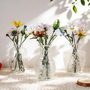 May include: Three clear glass vases, each holding colorful artificial flowers. The vases have different designs: ribbed, patterned, and textured. The flowers include yellow, white, pink, and blue blooms, creating a vibrant display.