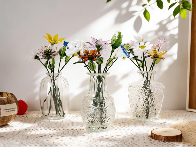 May include: Three clear glass vases, each holding colorful artificial flowers. The vases have different designs: ribbed, patterned, and textured. The flowers include yellow, white, pink, and blue blooms, creating a vibrant display.