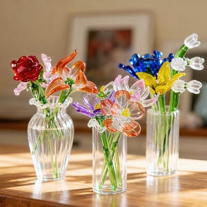 Op de afbeelding: Drie glazen vazen met kleurrijke glazen bloemen op een houten tafel. De bloemen zijn onder meer een rode roos, oranje lelies en blauwe, gele en witte bloesems. De vazen zijn helder en cilindrisch, met een geribbeld ontwerp.