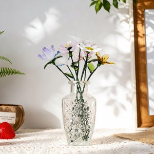 May include: A clear glass vase with a geometric pattern holds a bouquet of colorful glass flowers. The flowers include purple, pink, white, and yellow blooms with green stems and leaves. The vase sits on a white lace tablecloth.