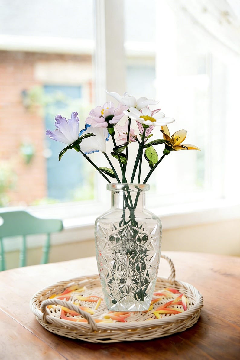 May include: A clear glass vase with a geometric pattern holds a bouquet of colorful glass flowers. The flowers include white, pink, yellow, and blue petals with green stems. The vase sits on a woven tray with a decorative border.