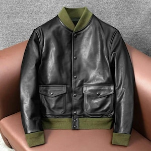 Peut inclure: Veste de bombardier en cuir noir avec col, poignets et ourlet verts. La veste a deux poches avant et une fermeture boutonnée. La veste est en cuir lisse et brillant.